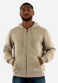 Sudadera con cremallera en color beige, hecha de tela suave, con capucha forrada, cordones y bolsillos frontales. Destacada con un parche de logo en la parte inferior izquierda.