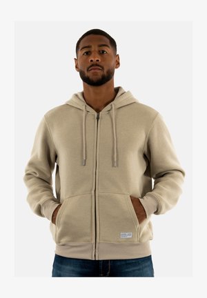 Sudadera con cremallera en color beige, hecha de tela suave, con capucha forrada, cordones y bolsillos frontales. Destacada con un parche de logo en la parte inferior izquierda.