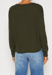 Haut à manches longues côtelé vert foncé avec un col arrondi ; coupe ample, texture douce et manches à poignets, associé à un jean en denim bleu clair.