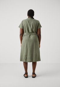 Oliwkowa sukienka do kolan z krótkim rękawem, pasek w talii i prosty krój, wykonana z lekkiego materiału. Na nogach czarne płaskie sandały.