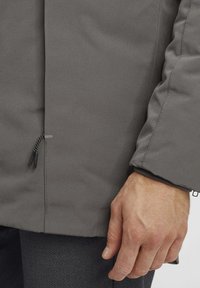 Chaqueta gris texturizada con un bolsillo con cremallera, puños elásticos y una silueta aerodinámica, con un primer plano de una mano agarrando la tela.