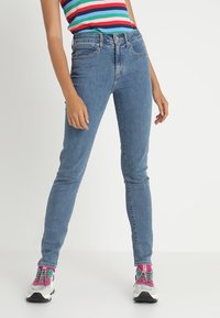 Jeans ajustados de color azul hechos de mezclilla, con una cintura de altura media, bolsillos frontales y un corte slim que llega hasta el tobillo. Llevados con zapatillas multicolores.
