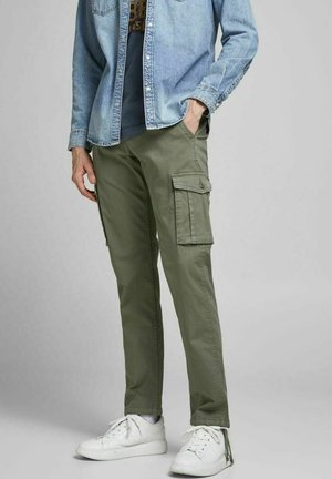 Hombre con pantalones cargo verde oliva, zapatillas blancas y una camisa vaquera azul claro con una mano en el bolsillo.