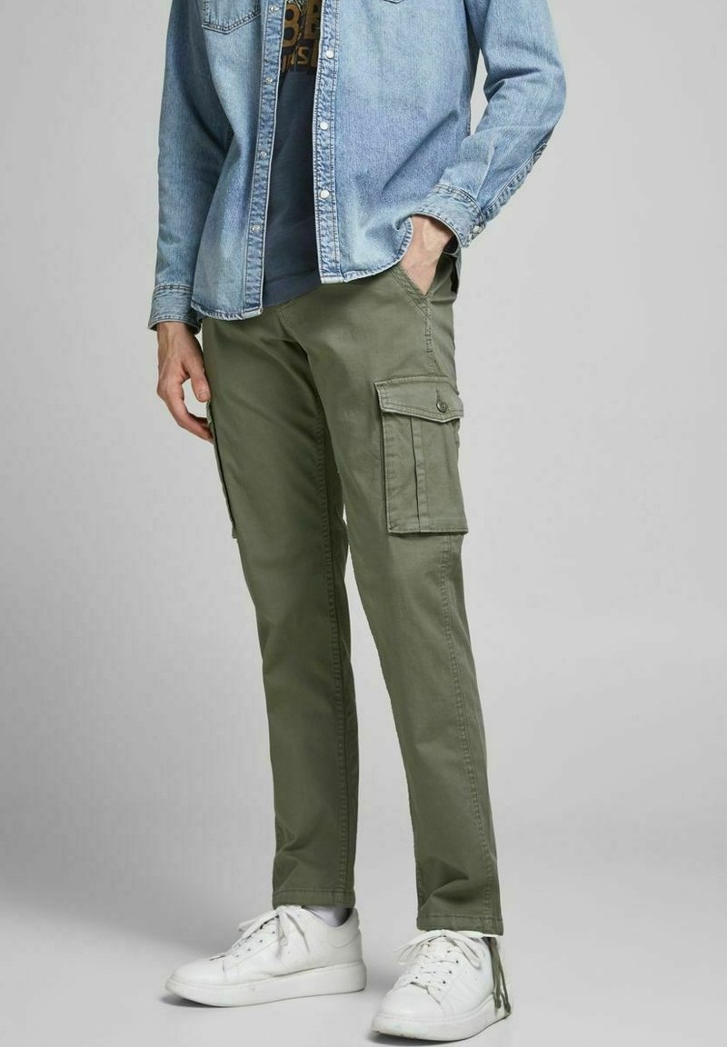 Homme portant un pantalon cargo vert olive, des baskets blanches et une chemise en jean bleu clair avec une main dans la poche.