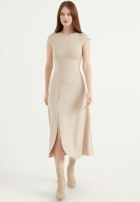 Beige, tailliertes Midi-Kleid mit kurzen Ärmeln, das einen Schlitz vorne und eine glatte Textur aufweist. Getragen mit passenden Stiefeln bis zum Knöchel.