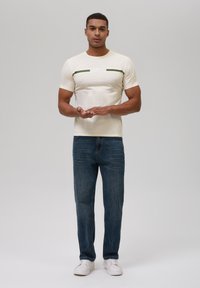 Camiseta ajustada beige con una franja verde y un logo en el pecho, combinada con jeans rectos azules y zapatillas blancas. Diseño sencillo.