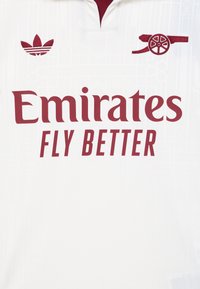 Bijela nogometna majica s ovratnikom. Sadrži crveni vezeni natpis "Emirates Fly Better" i logo, uz crvenu Adidas Trefoil oznaku.