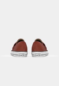 Tênis baixos de camurça vermelha com solados de borracha branca e acabamentos em listras preto e branco. Apresenta a marca Converse All Star no calcanhar.
