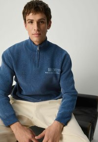 Mėlynas, pūkuotas pusiau užtraukiamas megztinis su raukšlėta apykakle ir rankogaliais; ant viršaus balta spalva užrašyta "BROOKLYN WILLIAMSBURG". Paprastos šviesios spalvos kelnės.