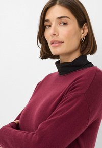 Maglione lavorato a maglia color bordeaux con maniche lunghe e scollo rotondo, sovrapposto a un dolcevita nero. Presenta una texture morbida e una vestibilità comoda.