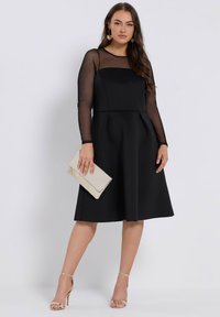 Nainen, jolla on päällään musta mekko, jossa on läpikuultavat pitkät hihat. Hän pitää kädessään kultasilkkistä laukkua ja hänen jaloissaan on kultaiset korkokengät.