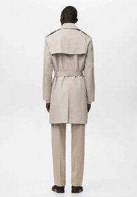 Beżowy trencz z paskiem w talii, rozcięciem z tyłu i pagonami na ramionach, noszony z dopasowanymi spodniami i czarnymi butami.