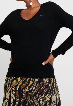 Personne portant un pull noir col en V avec un petit logo sur la poitrine et une jupe à motifs avec un design abstrait marron, noir et beige.