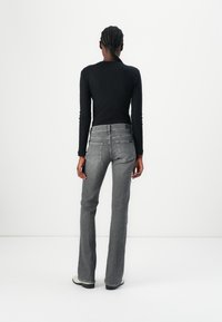 Personne aux cheveux tressés, vue de dos, portant un haut noir à manches longues, un jean gris et des chaussures blanches sur un fond blanc uni.