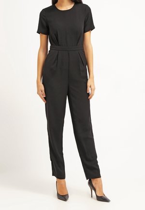 Woman wearing a black short-sleeve jumpsuit with a fitted waist and black pointed-toe high heels, standing against a white background.