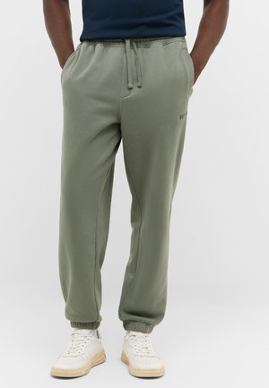 Green sweatpants with elastic cuffs, drawstring waist, side pockets, and subtle branding on the leg. Paired with white sneakers.