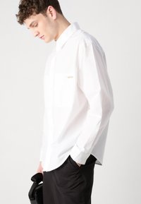 Biele button-up košeľa s vreckom a logom Calvin Klein; hladká textúra a dlhé rukávy. Skombinovaná s tmavými nohavicami, držanými čiernym predmetom.