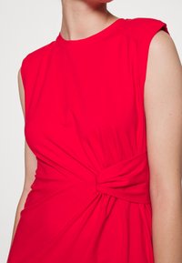 Röd klänning med korta ärmar och knutdetalj i midjan, tillverkad av mjukt och stretchigt tyg. Den släta texturen och den åtsittande designen framh�äver silhuetten.