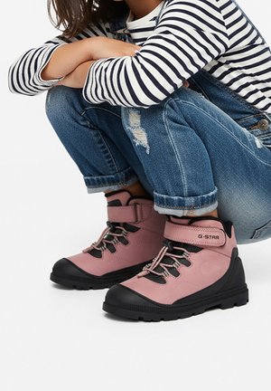 Bambino con jeans strappati arrotolati, maglia a maniche lunghe a righe e sneakers alte rosa con suole nere, seduto a gambe incrociate.