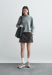 Pull à col roulé gris, mini-jupe en cuir marron avec des coutures visibles, et baskets marron, portés avec des chaussettes blanches mi-mollet et un sac noir.