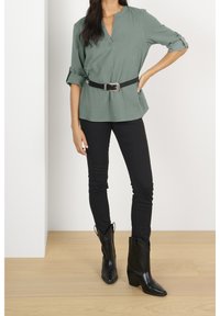 Top verde a maniche corte con scollatura a V e una cintura nera; abbinato a pantaloni skinny neri e stivali da cowboy neri con punta appuntita.