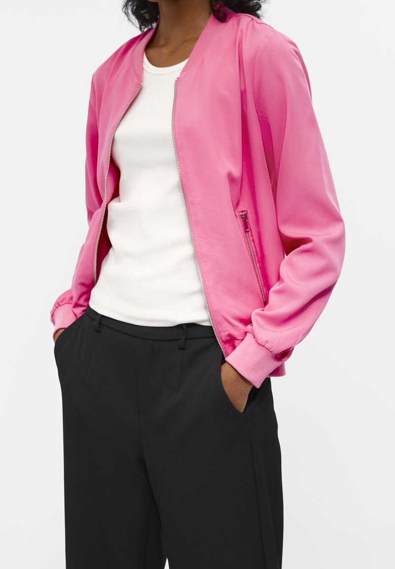 Femme portant une veste zippée rose vif sur une chemise blanche avec un pantalon noir, mains dans les poches, sur un fond uni.