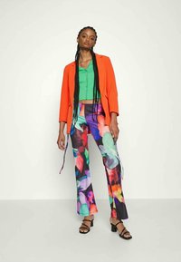 Bright orange blazer over a green top, paired with colorful patterned trousers and black heeled sandals. Multi-colored floral design on pants.