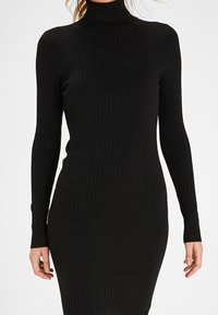 Robe à col roulé noire côtelée avec manches longues, coupe ajustée et tissu texturé, atteignant mi-mollet.