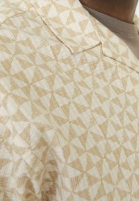 Kurzarmhemd aus leichtem beigem Stoff, mit einem geometrischen dreieckigen Muster in hellbraun, Kragen und strukturierten Details.