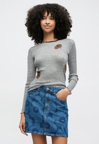 Göndör hajú nő szürke, hosszú ujjú felsőben, barna virágmintákkal, és kék farmer szoknyában, virágmintákkal, kezét a zsebében tartva.