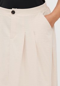 Φούστα πλισέ σε ανοιχτό μπεζ χρώμα φτιαγμένη από υφασμένο ύφασμα, με ζώνη που έχει ελαστική πλάτη και μπροστινή τσέπη. Λεπτομέρεια με ένα μόνο σκοτεινό κουμπί.