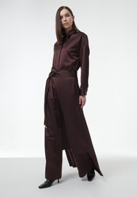 Vestido largo de satén marrón con cuello, cintura ceñida y aberturas laterales, combinado con tacones negros de punta. Textura suave, corte ajustado.