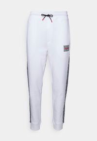 Weiße Jogginghosen mit elastischem Bund, schwarzem und rotem Kordelzug, Seitentaschen und kontrastierenden schwarzen gemusterten Seiteneinsätzen. Kleines Logo auf der Vorderseite.