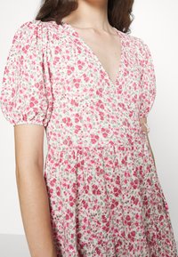 Mujer vestida con un vestido blanco con estampado floral rosa, de mangas cortas abullonadas y escote en V, mostrado de los hombros a la cintura.