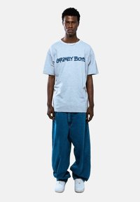 Szary t-shirt z krótkim rękawem z napisem "GRIMEY BOYS" w granatowym kolorze, zestawiony z luźnymi niebieskimi dżinsami i białymi trampkami.