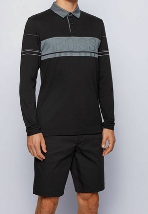 Man draagt een zwart poloshirt met lange mouwen, een grijze kraag, een grijze horizontale streep en een "BOSS" logo, gecombineerd met zwarte korte broek.