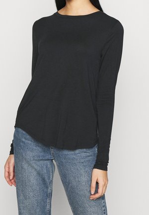 Schwarzes Langarmshirt mit rundem Ausschnitt und weicher Textur, das eine lockere Passform und einen leicht geschwungenen Saum aufweist. Kombiniert mit blauen Jeans.