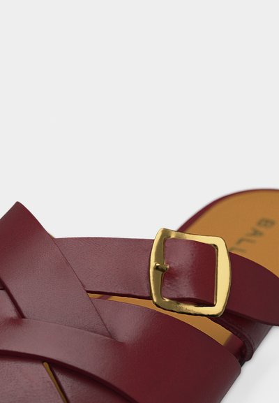 Gros plan sur une bride de sandale en cuir bordeaux avec une boucle carrée en or et une semelle intérieure beige.