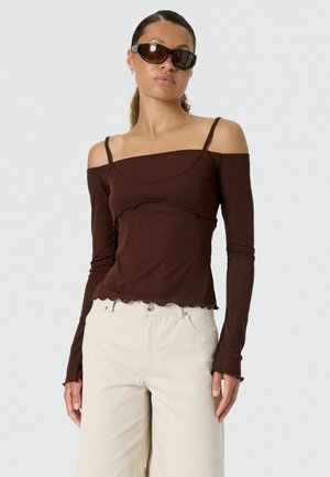 Vrouw draagt een donkerbruin off-shoulder top met lange mouwen en dunne bandjes, lichtbeige broek en donkere zonnebril tegen een effen achtergrond.