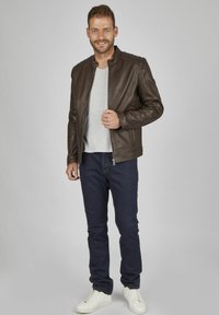 Braune Lederjacke mit Stehkragen, Reißverschluss und Seitentaschen, kombiniert mit einem grauen T-Shirt, dunklen Jeans und weißen Sneakern.