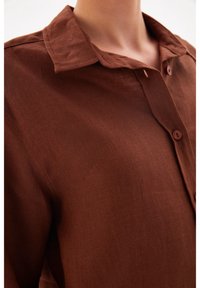 Brown linen shirt featuring a button-up front, a classic collar, and visible texture with a relaxed fit. Accented by matching buttons.