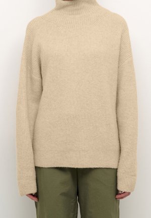 Beige ribstrikket rullekravetrøje med lange ærmer, kombineret med løse olivengrønne bukser på en stående person.