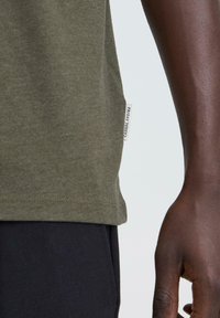 Maslinasto zelena pamučna majica kratkih rukava, sa zaobljenim rubom i bočnom oznakom s natpisom "FRIDAY" blizu dna. Glatka tekstura.