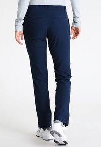 Mörkblå byxor i flexibelt tyg, med smal passform, två bakfickor och en mjuk yta. Bärs med vita sportskor.