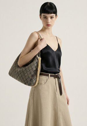 Donna con capelli corti e scuri che indossa una canotta in raso nero e una gonna beige, che tiene una grande borsa tote con motivo e tracolla a catena.
