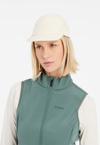 Světlé hnědé kšiltovka s zakřiveným kšiltem, vyrobená z měkkého materiálu. Nosí se s zelenou vestou na zip s vysokým límcem a detailem loga.