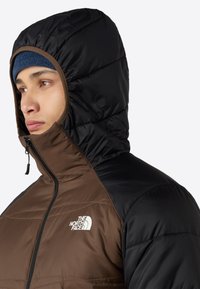 Braune und schwarze Pufferjacke mit Kapuze, ausgestattet mit einem Reißverschluss und dem Logo von The North Face auf der Brust. Glatte Textur, gesteppter Design.