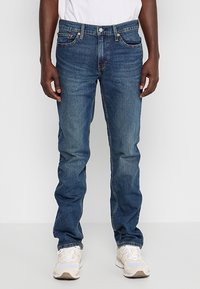 Homme portant un jean droit bleu avec des baskets blanches et une chemise blanche, debout devant un fond clair uni.