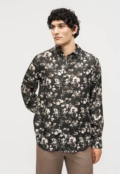 Άνδρας με σγουρά μαλλιά που φοράει μαύρο πουκάμισο με λουλούδια και κουμπιά και ανοικτό καφέ παντελόνι, κοιτάζοντας στο πλάι σε απλό φόντο.