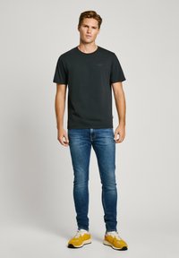 Camiseta de algodón negra con cuello redondo y mangas cortas, combinada con jeans de mezclilla azules y zapatillas amarillas y blancas. Modelo en posición erguida.